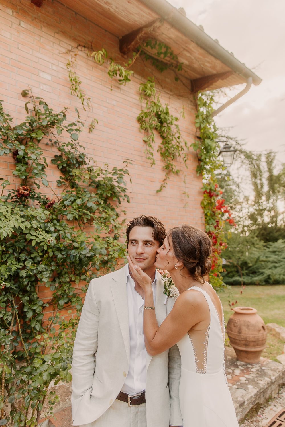 A couple in elegant white wedding attire share an intimate moment by a rustic wall with climbing roses.