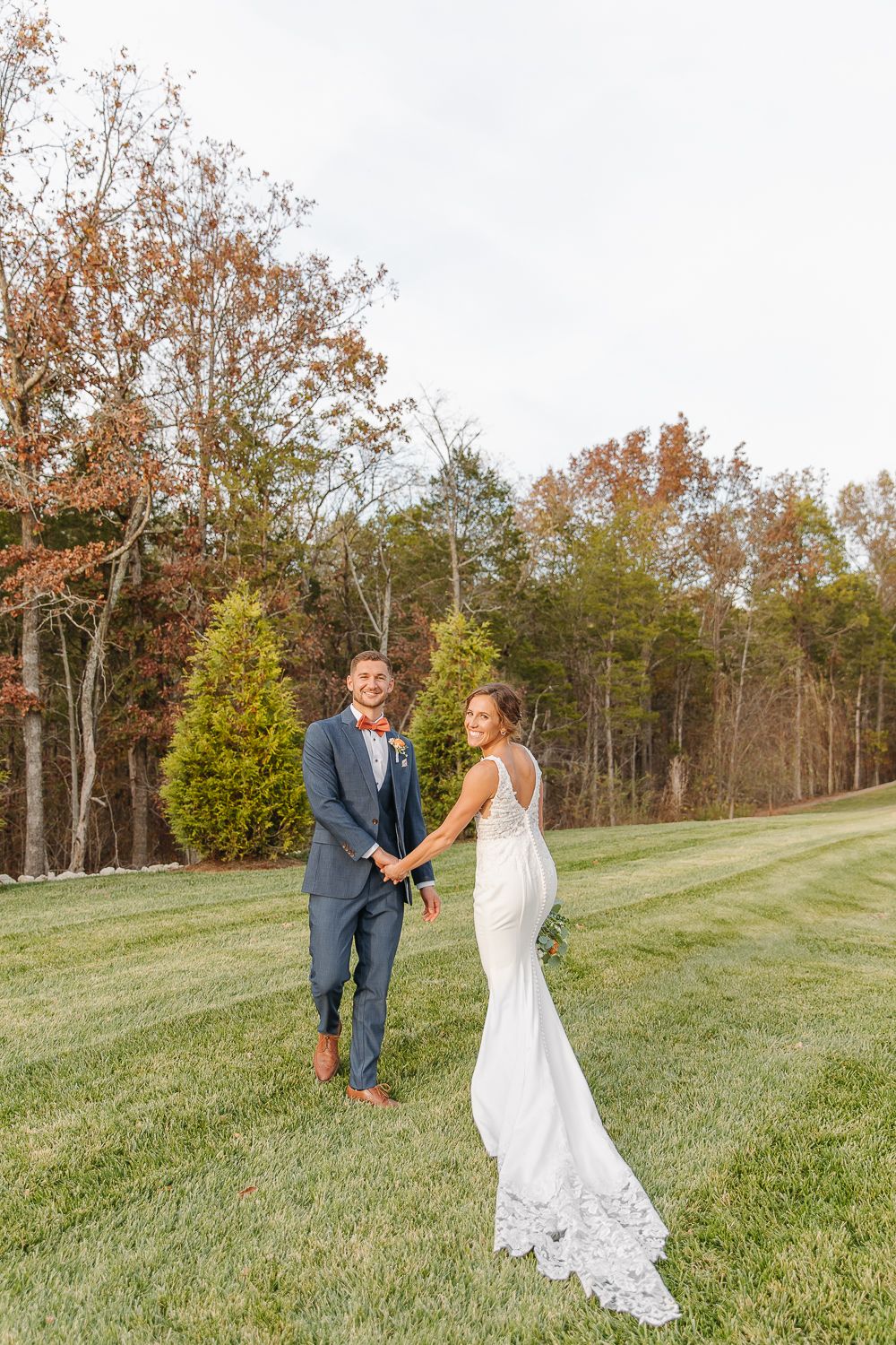 Couple holding hands in a fitted suit and white lace mermaid wedding dress on a grassy hillside during autumn.