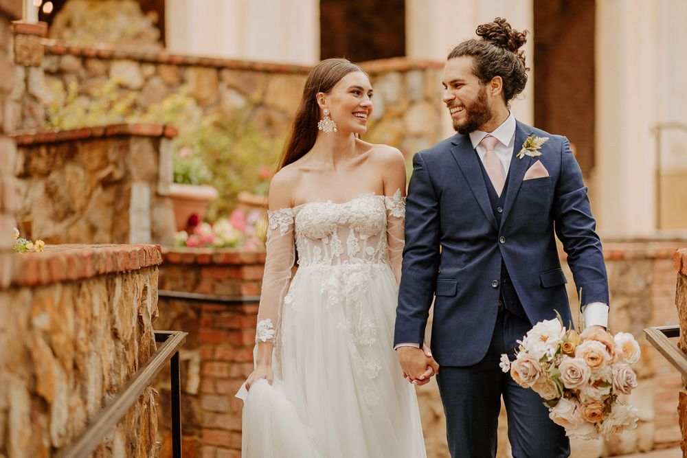 A couple in wedding attire walks down stone steps with the bride holding a peach and white floral bouquet in at the Bella Collina Venue.