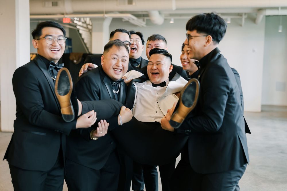 Groomsmen in black suits laugh and joke together during wedding party celebration.