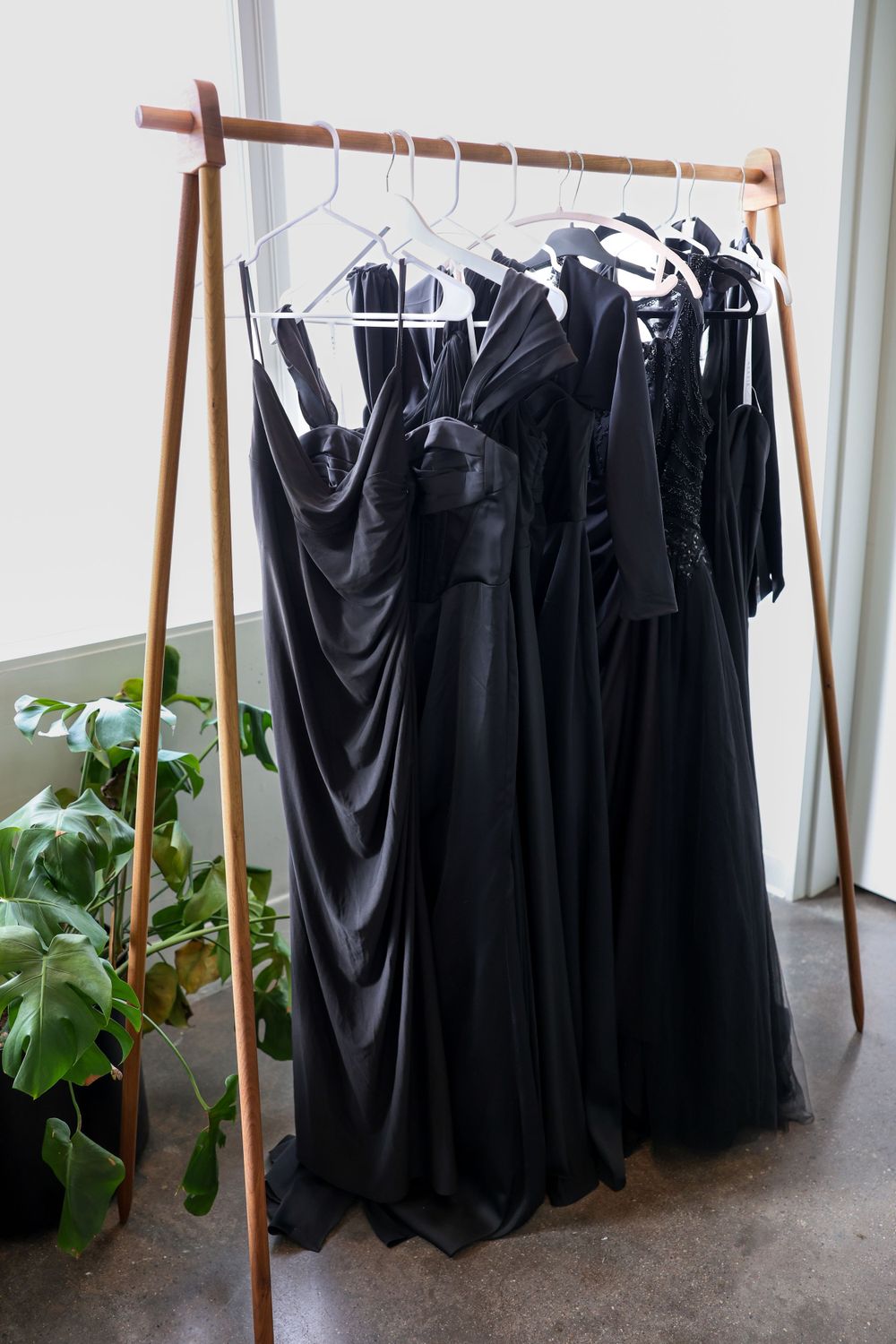 Black formal dresses hanging on a wooden clothing rack with a green potted plant beside it.