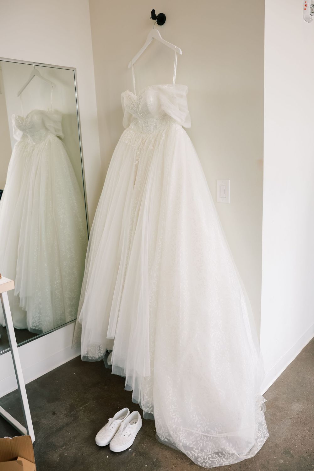 White tulle wedding dress with lace trim hangs beside a mirror with white ballet flats on the floor.