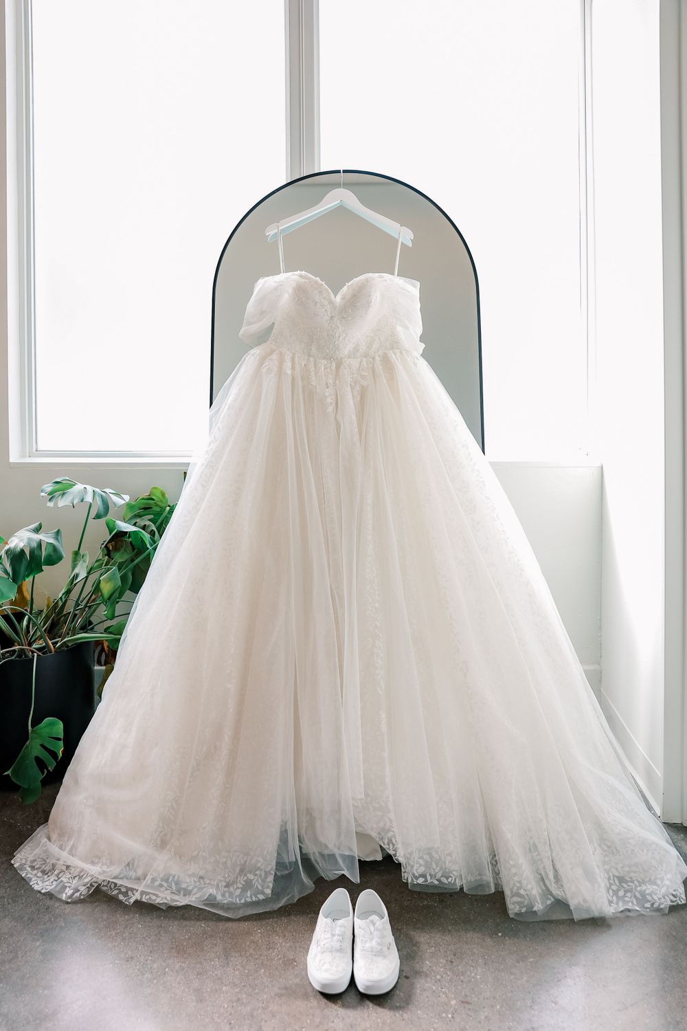 Elegant white strapless wedding gown with tulle skirt and lace detail hanging in window arch with matching white shoes below.