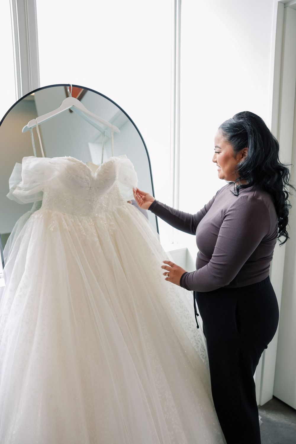 A person in a brown sweater adjusts a white strapless wedding gown hanging on display.