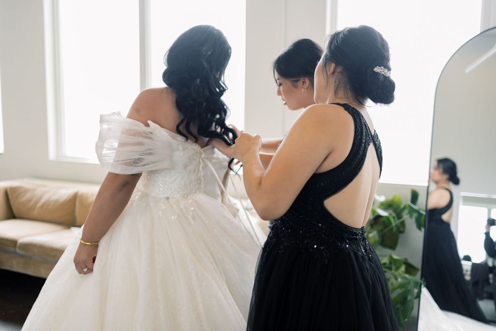 A bride in a white off-shoulder wedding gown gets help from someone in a black dress during wedding preparations.