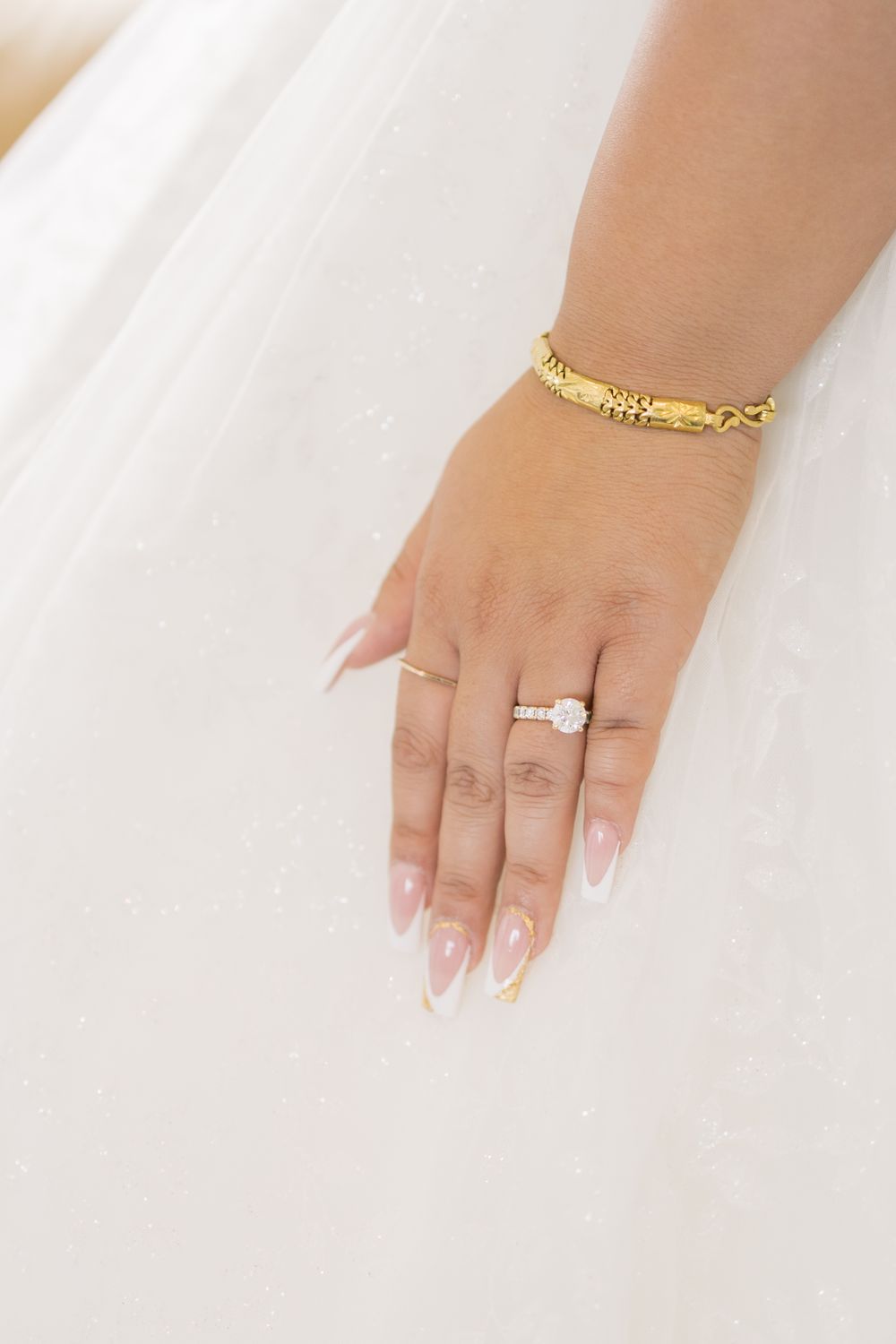 Hand with French manicured nails wearing an elegant gold bracelet and diamond engagement ring against white fabric.