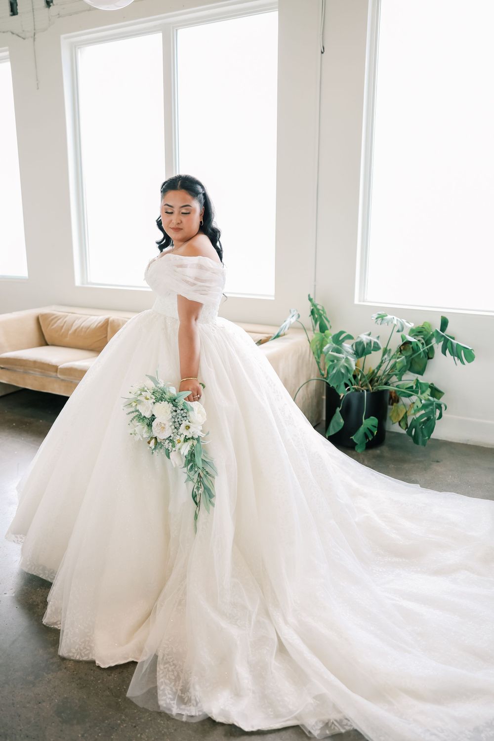A bride in an elegant off-shoulder white ballgown with long train holds a cascading green and white bouquet in a bright room.