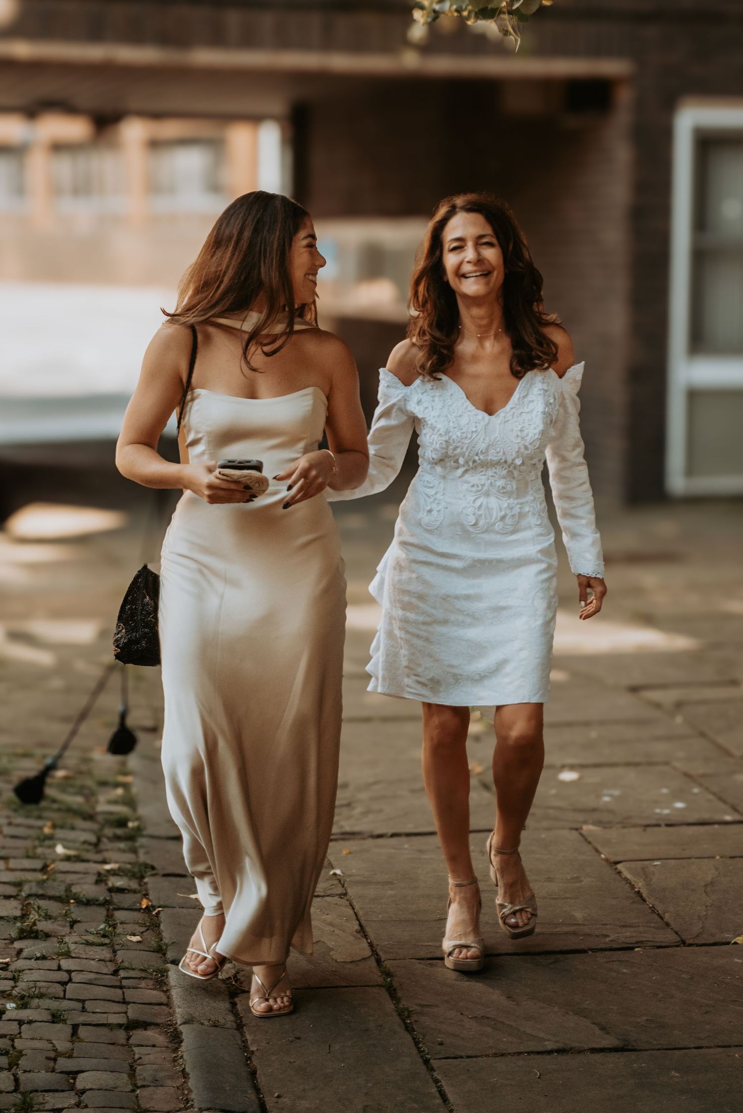 Two women in white dresses walking and laughing together on a cobblestone path during golden hour.
