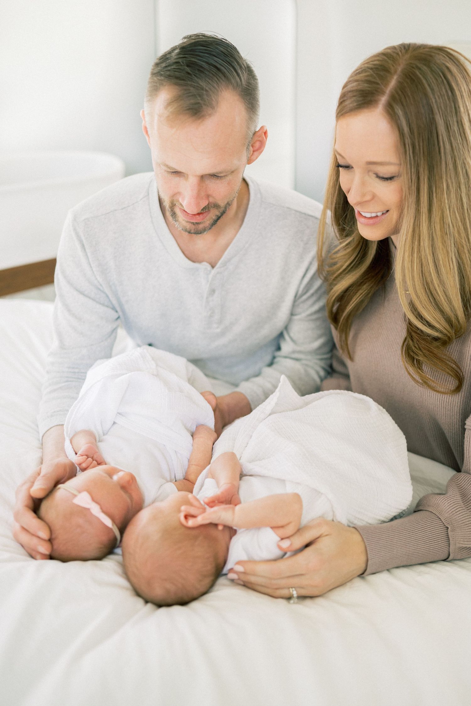 A tender moment with newborn twins and parents in white and beige clothing on a bed.