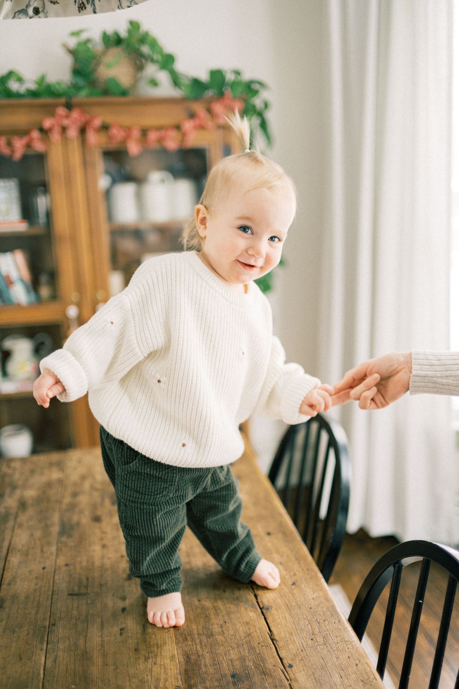 Baby in white sweater and corduroy pants standing on wooden table in cozy home setting.