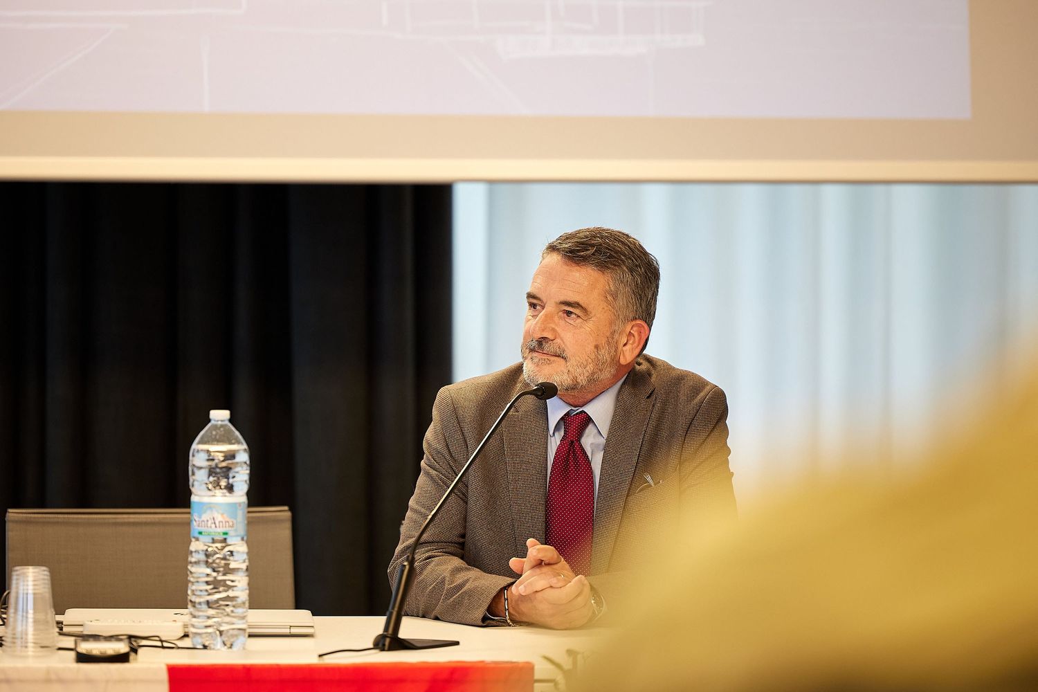 Speaker at conference table with microphone and water bottle.