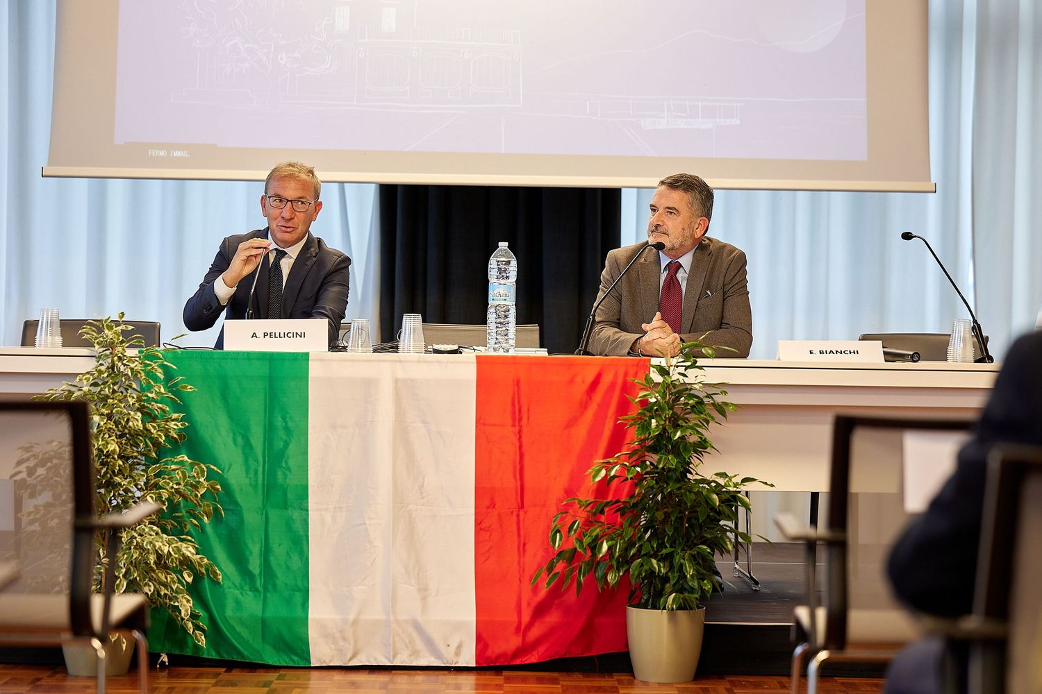 Panel discussion with two speakers seated at table with Italian flag decoration and potted plants.