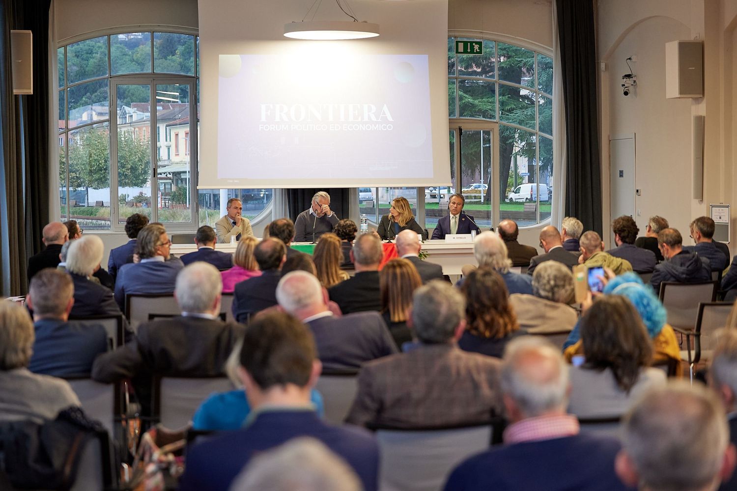 Large audience seated in conference room with projection screen and windows overlooking water.