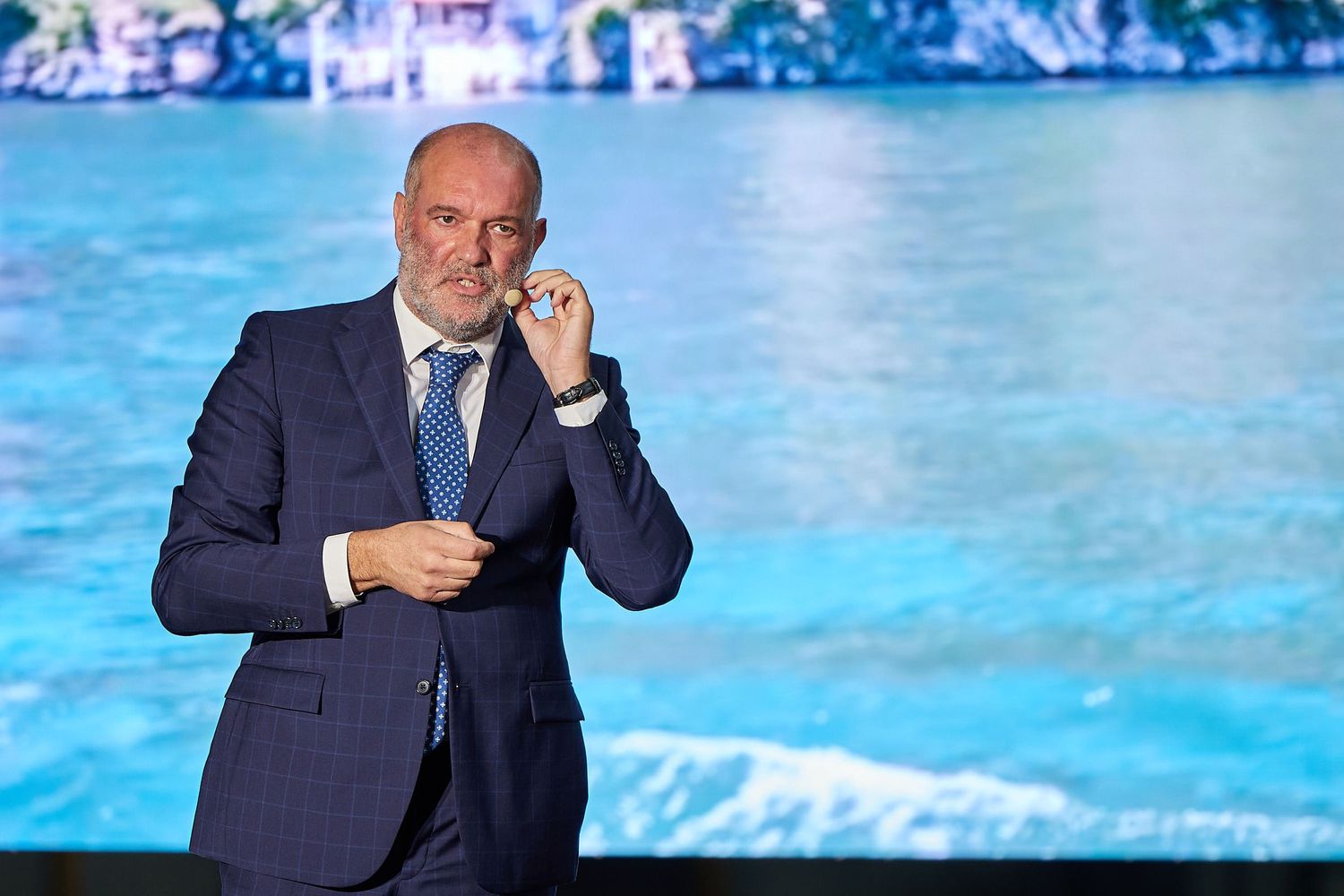 A businessman in a navy suit speaking on stage with a blue water backdrop.