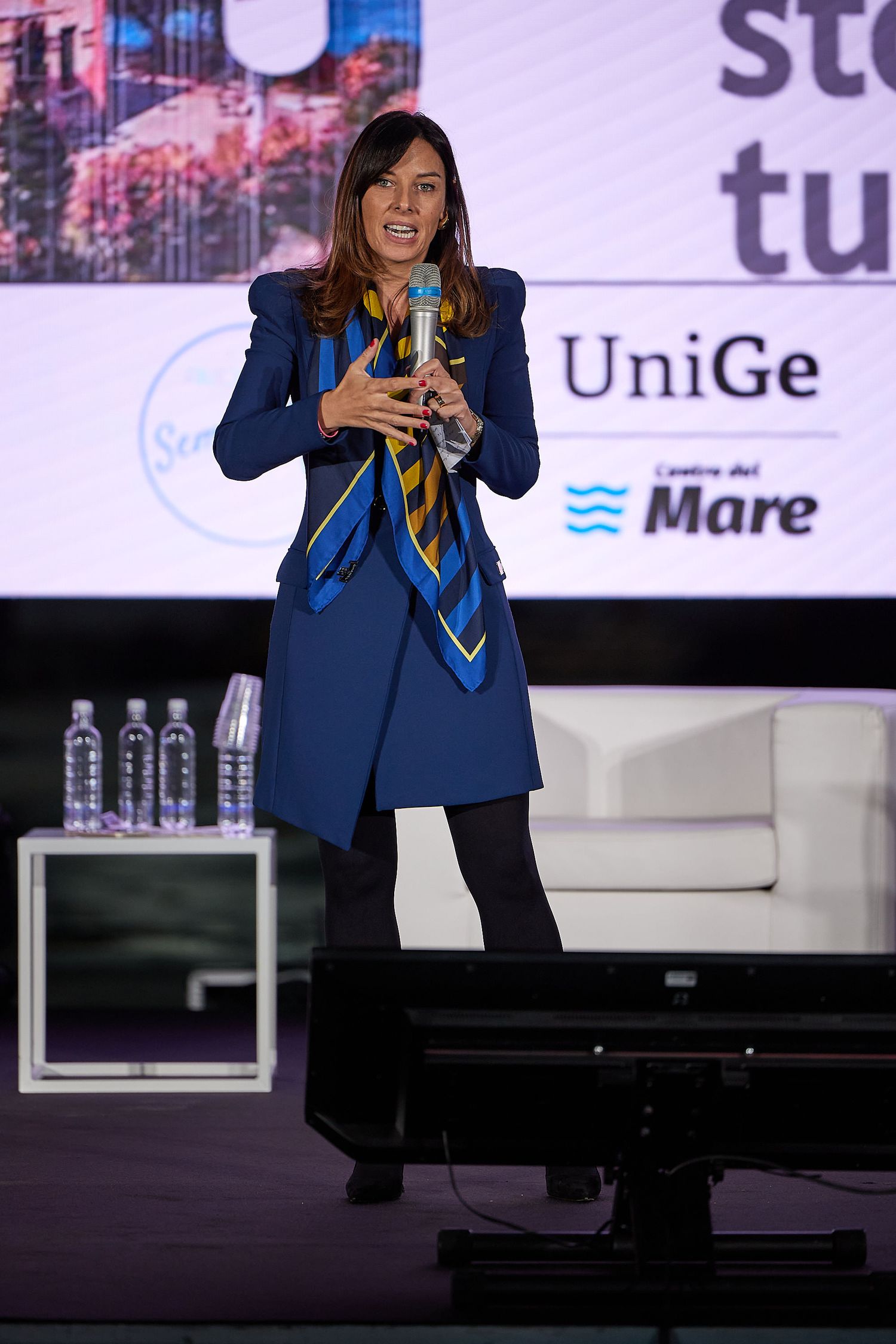 A presenter in a navy blue outfit speaking on stage with a branded backdrop.