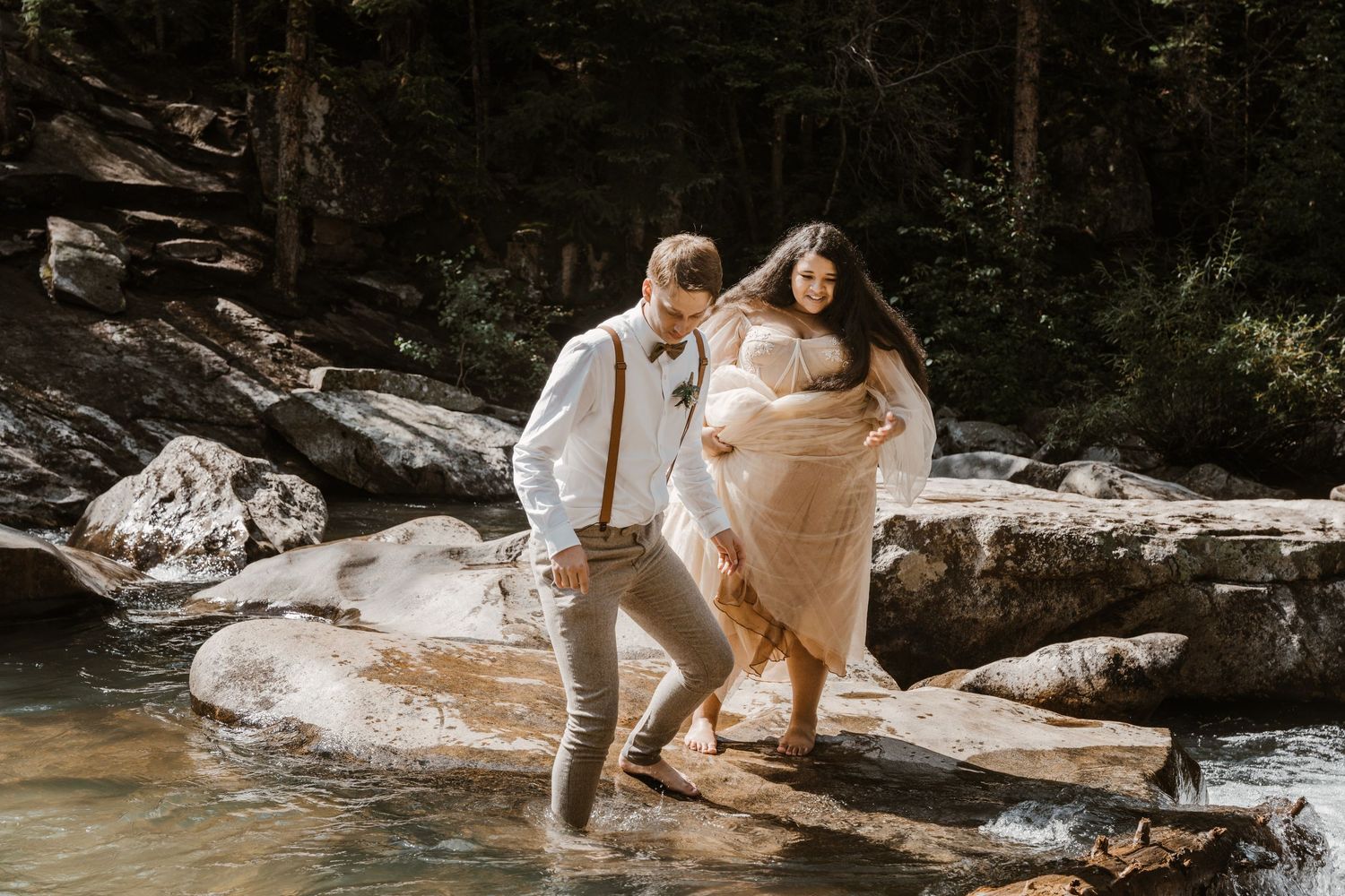 A couple stands together on rocky terrain near a stream in a forest setting.