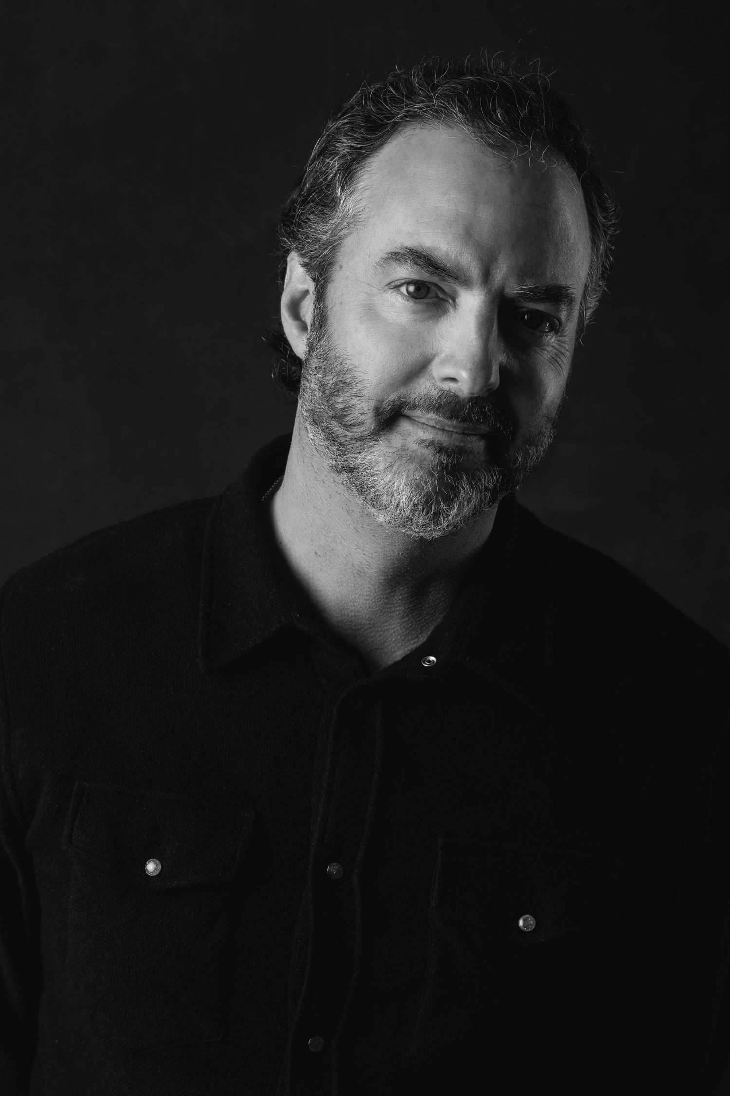A black and white studio portrait of a bearded man wearing a dark shirt against a dark background.