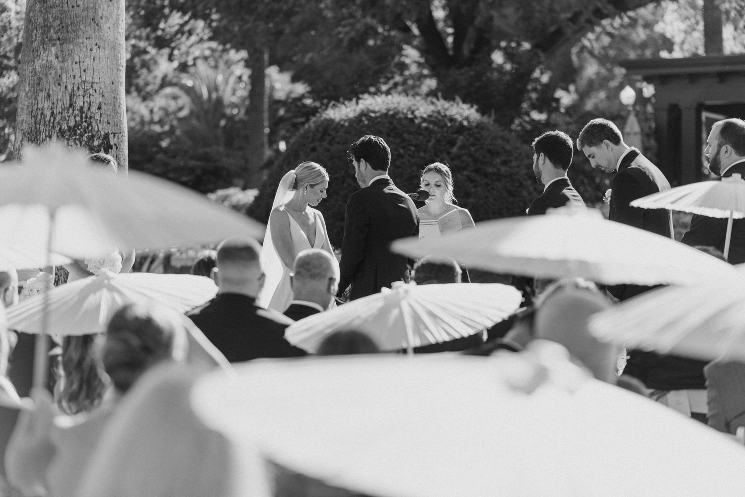 Wedding guests holding white parasols during an outdoor ceremony in black and white.