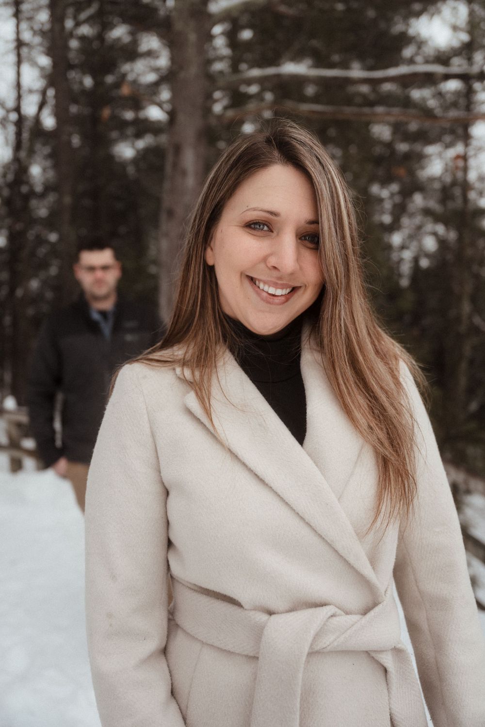 A person in a cream coat smiles while standing in a snowy forest setting.