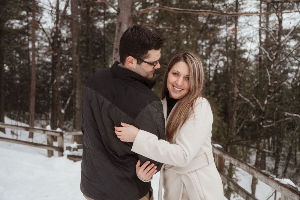 A couple embraces while standing in a snow-covered forest setting.
