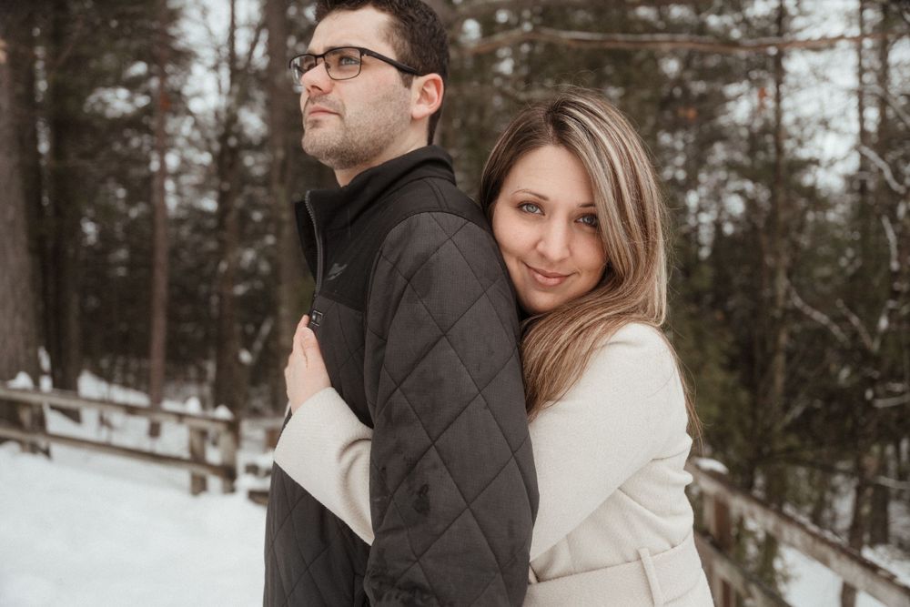 A couple poses together against a snowy forest backdrop wearing contrasting dark and light coats.