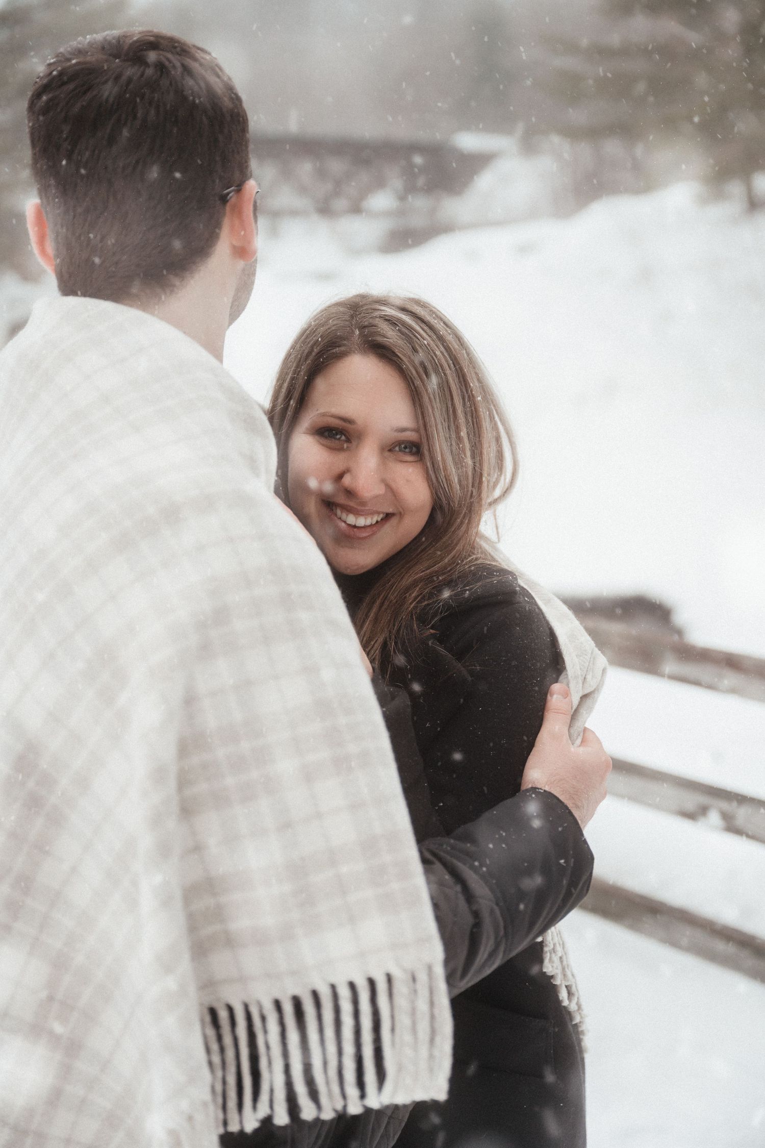 A couple shares a loving embrace in the snow while wrapped in a white fringed scarf.