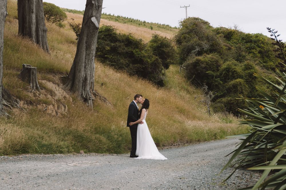 A romantic wedding couple embraces on a rural gravel road surrounded by trees.