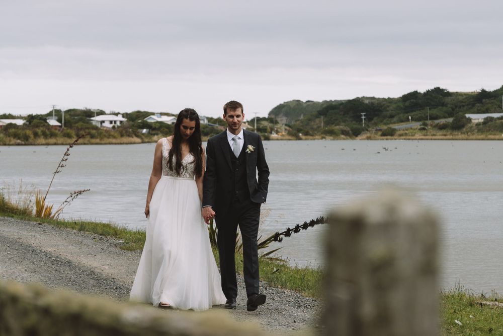 Wedding couple strolls along coastal path with water and houses in background.