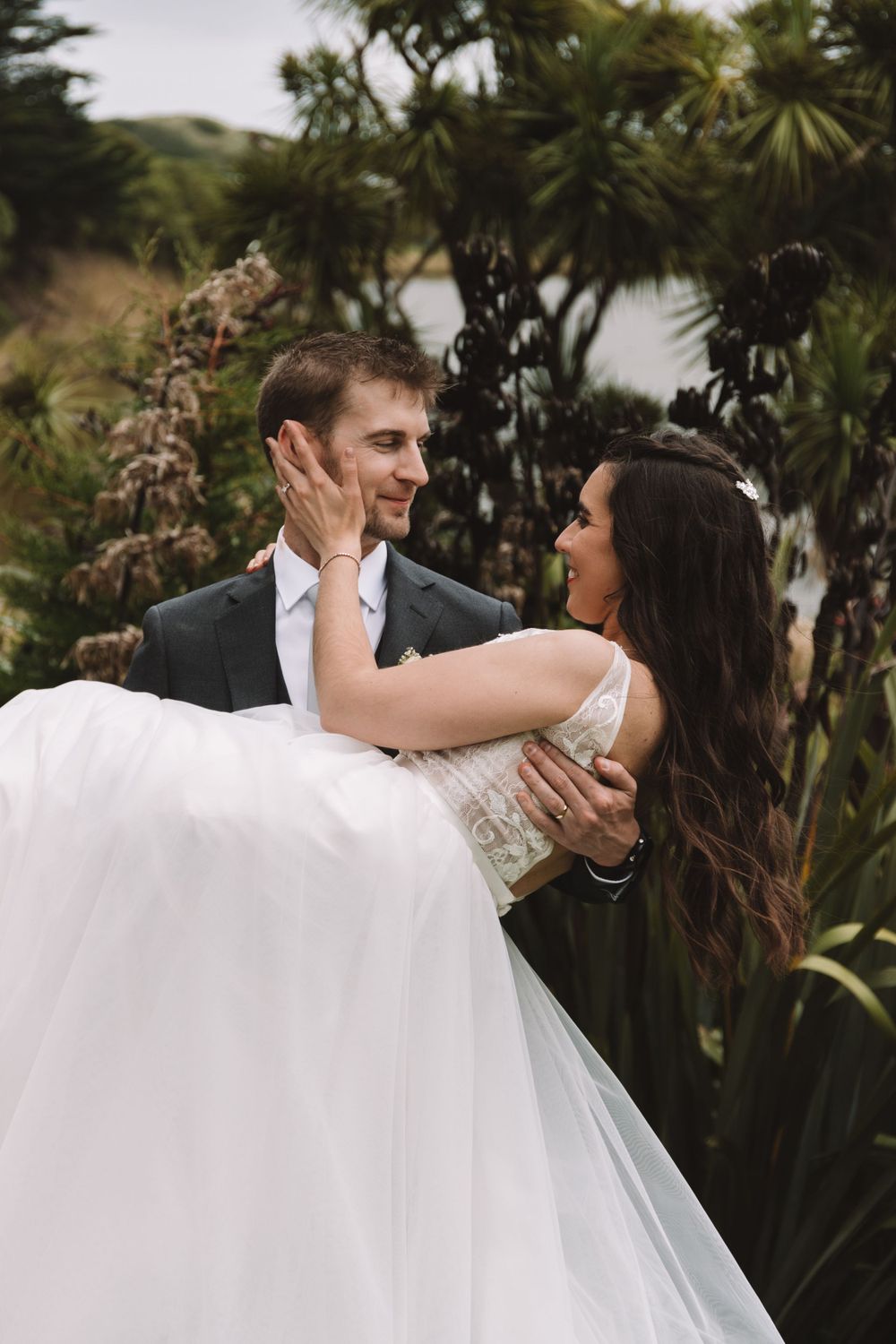 Groom carries bride in white dress among tropical foliage by the water.