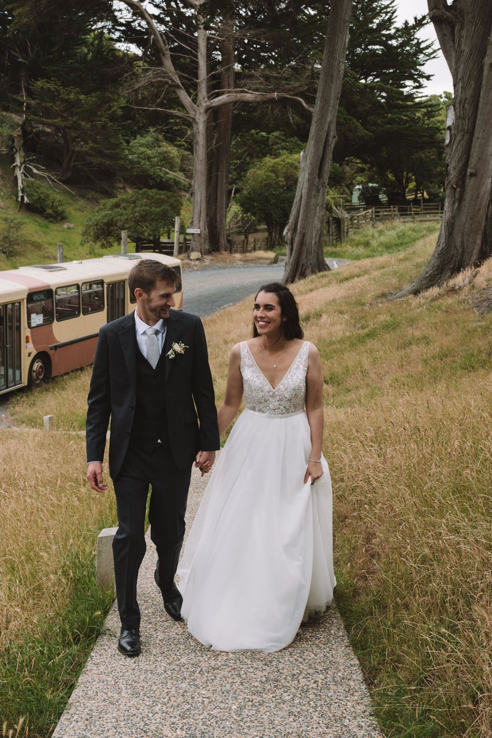 Wedding couple walking and laughing on path with vintage bus in background.