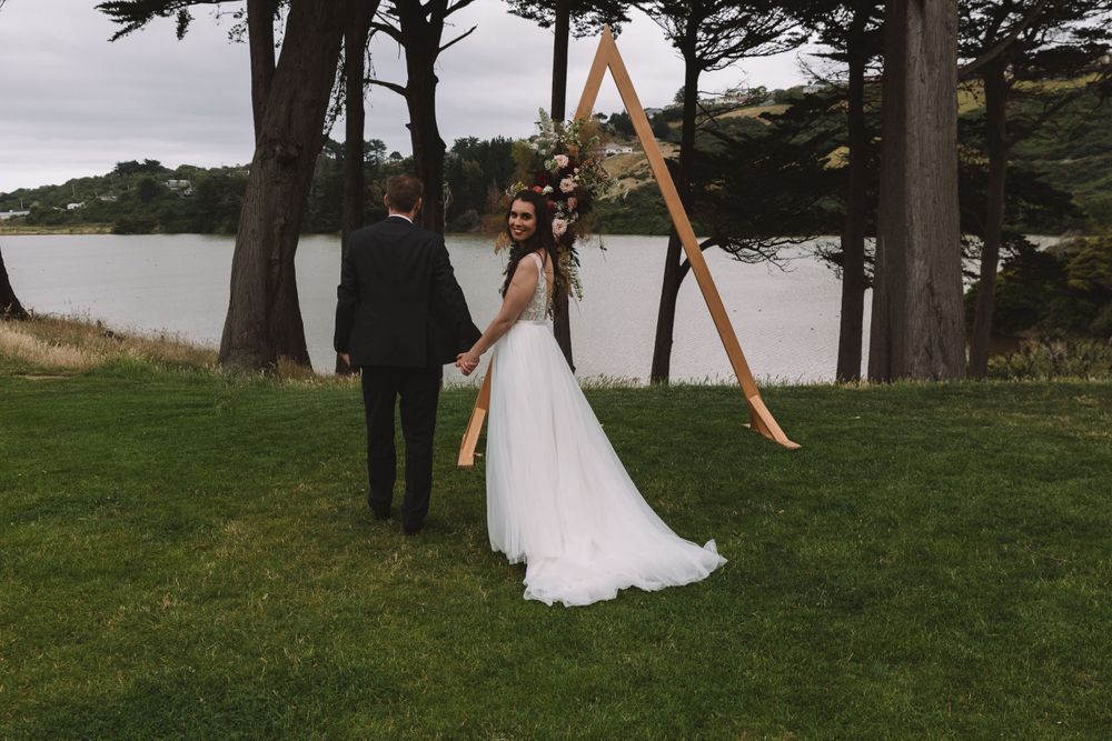 Wedding couple stands before wooden arch frame with lake view backdrop.