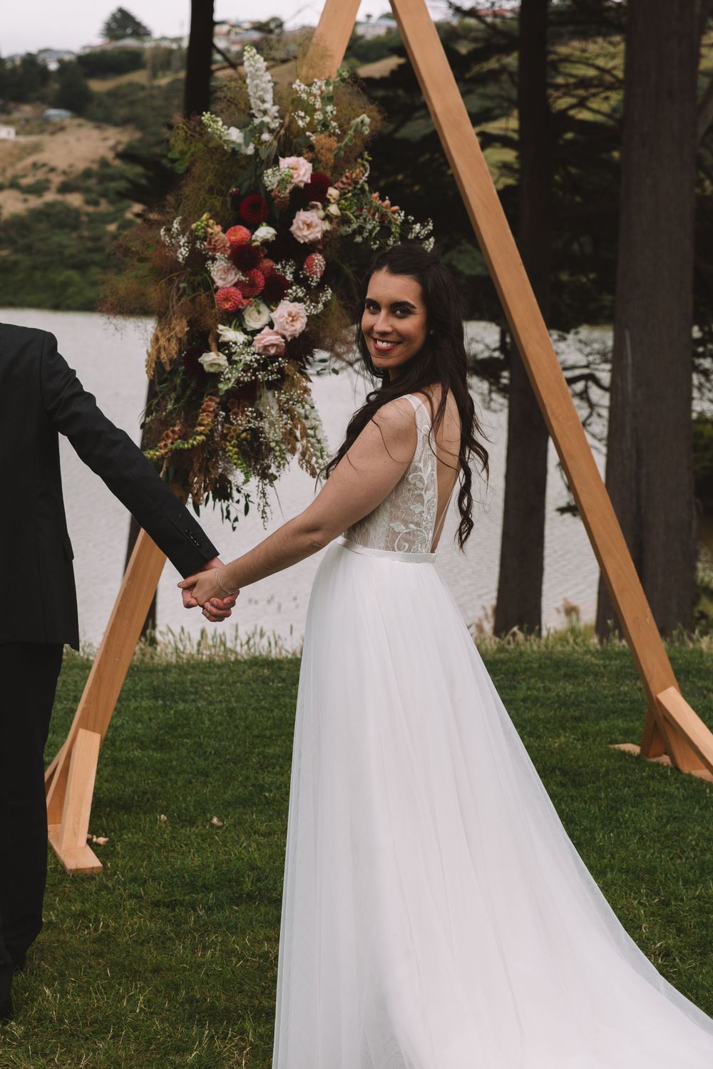 Bride poses by wooden triangle arch decorated with wild flowers and greenery.