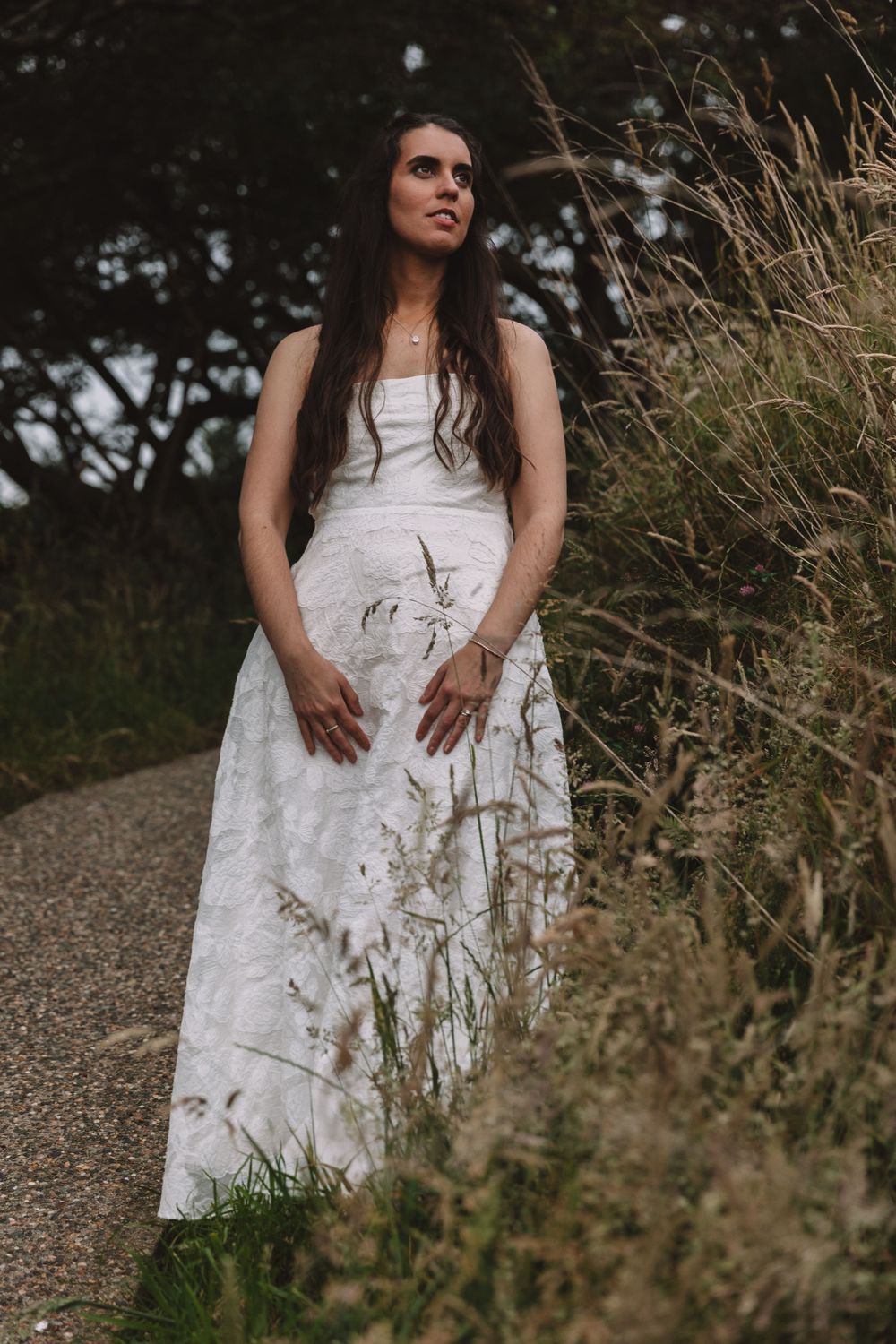 A bride in a lace dress poses thoughtfully among tall grasses on a nature path.