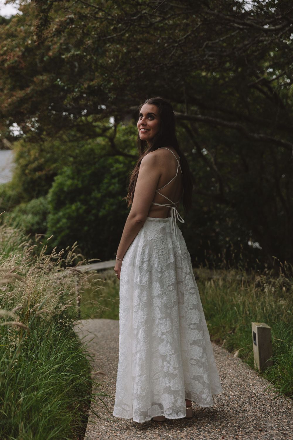 An elegant backless wedding gown is showcased against a natural outdoor backdrop with tall grasses.