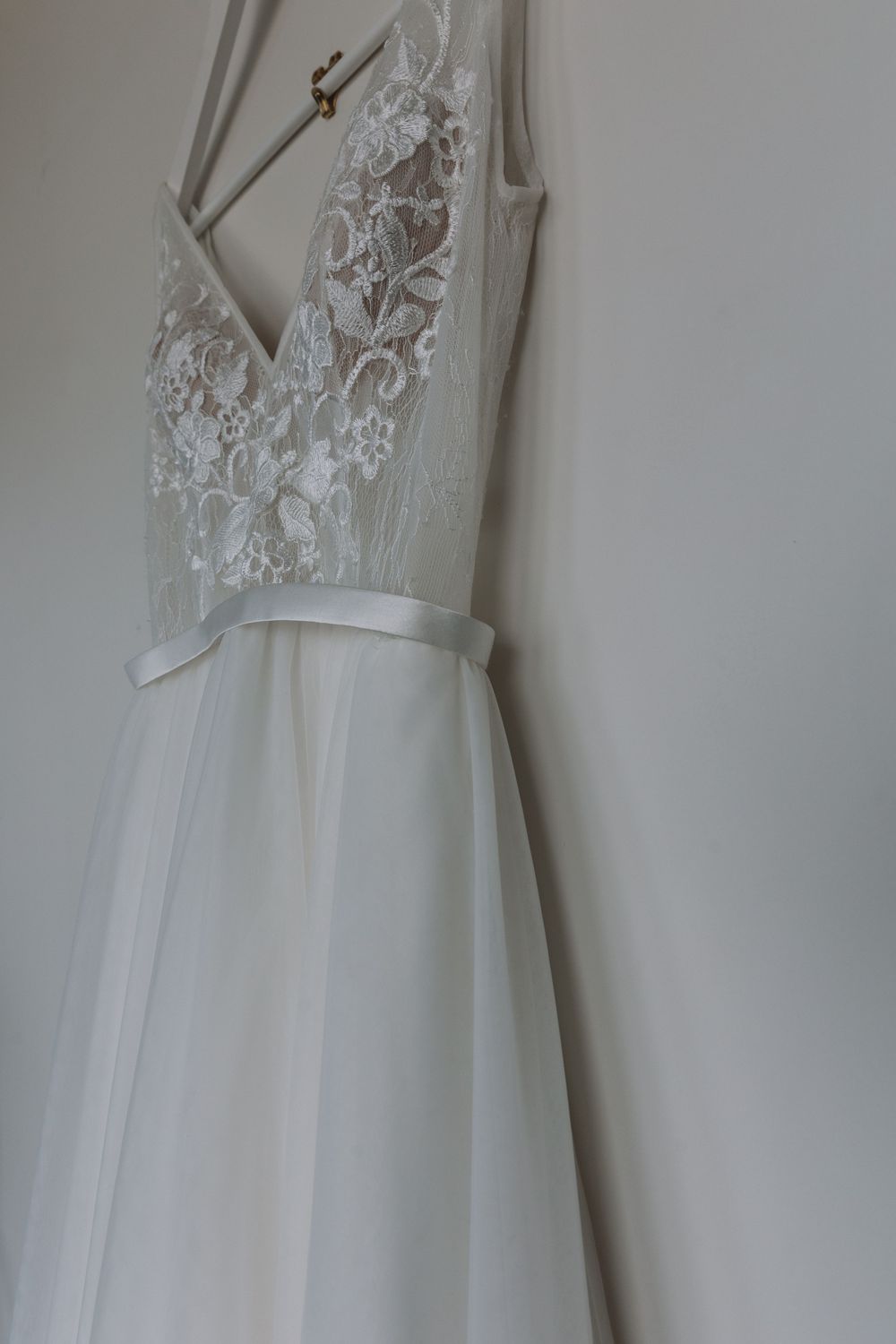 A white wedding dress with delicate lace bodice and flowy tulle skirt hangs elegantly against a light wall.