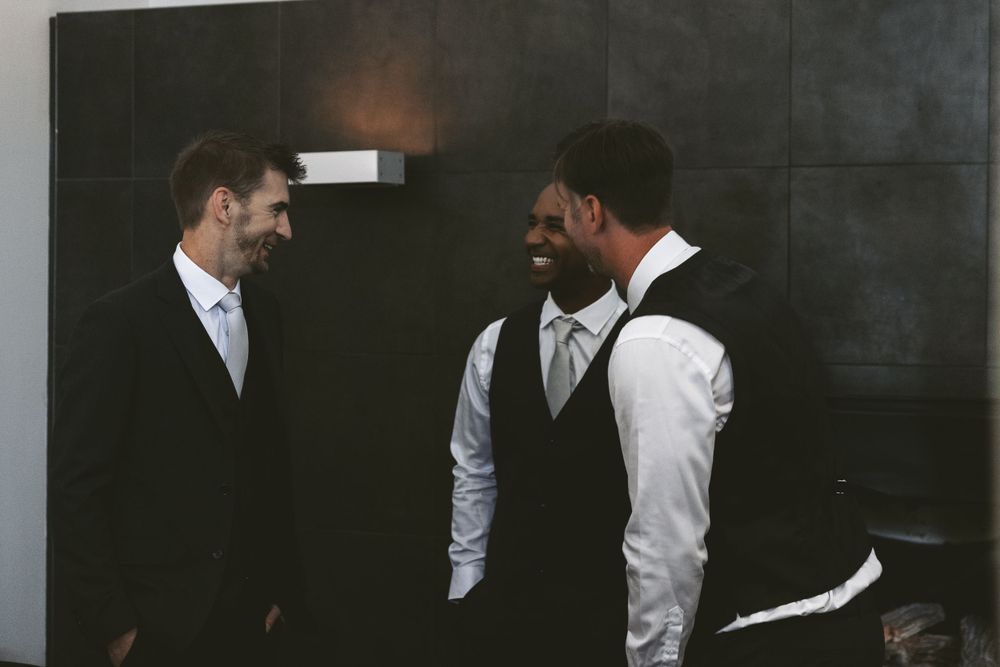 Two groomsmen in formal attire share a laugh in a dark hallway.