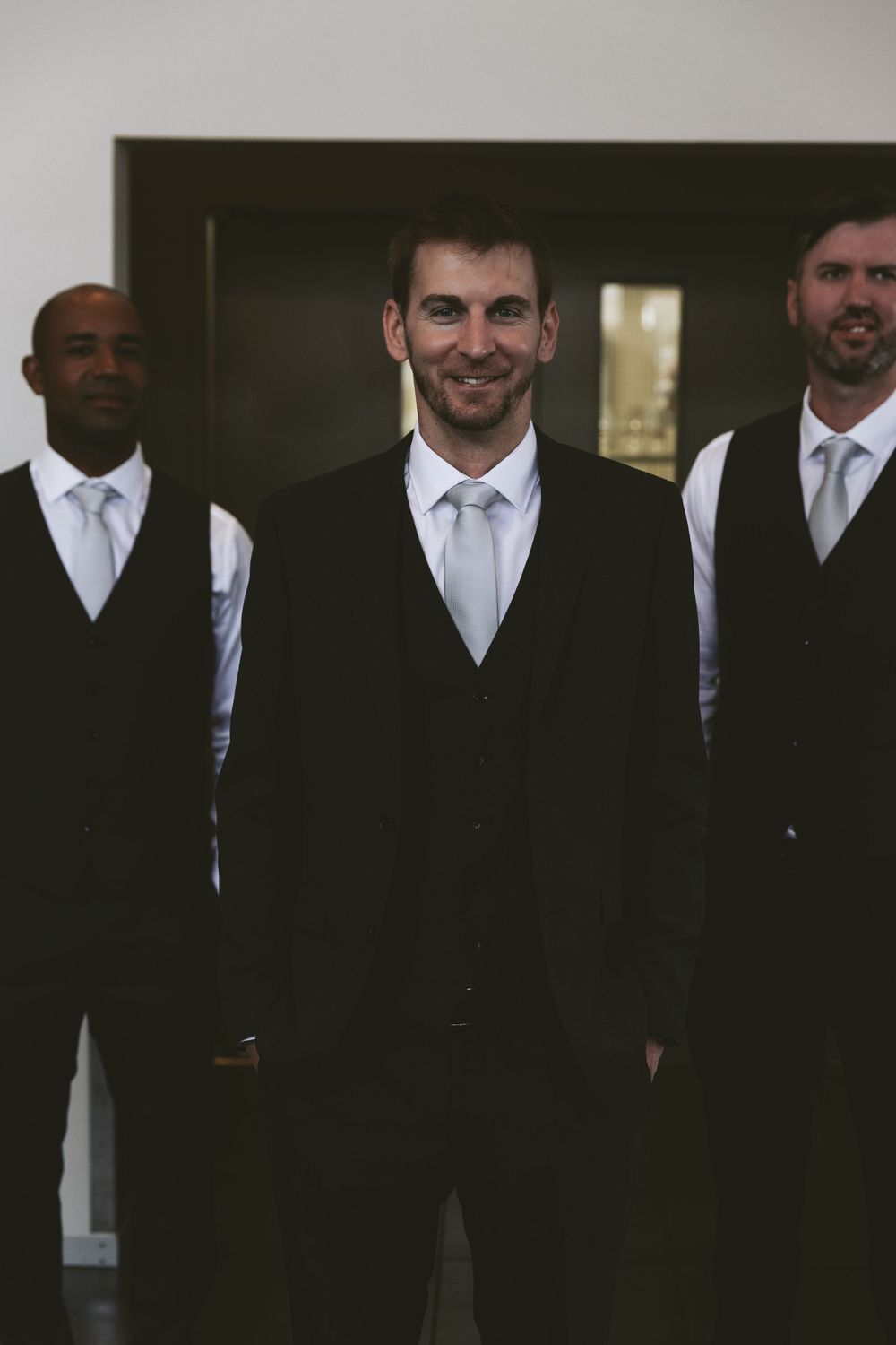 Three groomsmen in black suits and vests pose together for a formal wedding photo.