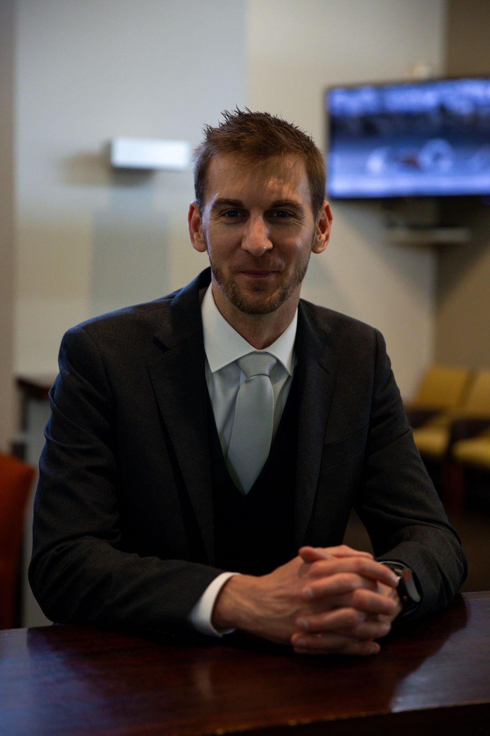A person in a dark suit and light tie sits at a wooden table with folded hands.