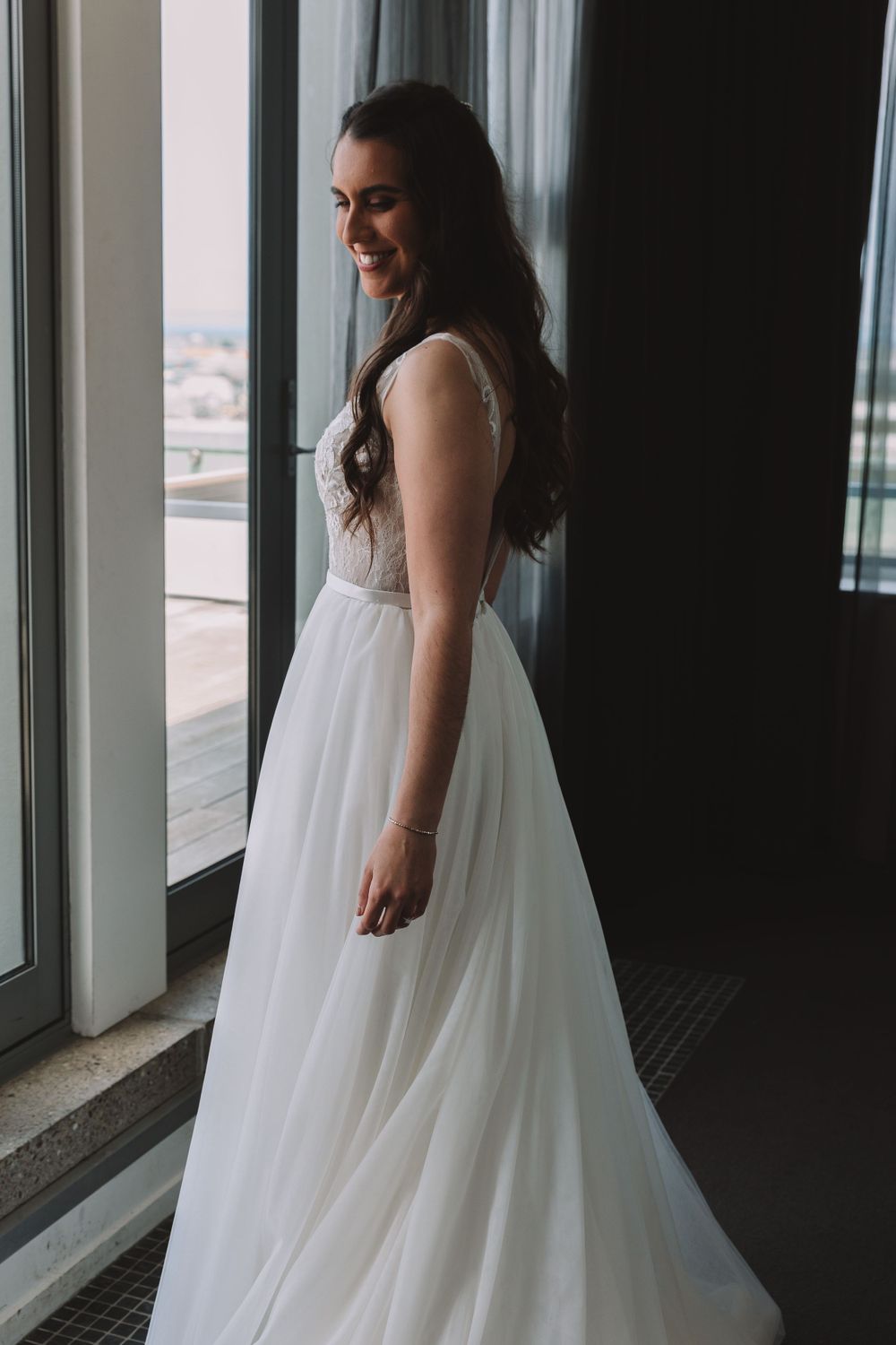 A bride in a flowing white wedding dress stands by floor-to-ceiling windows with city views.
