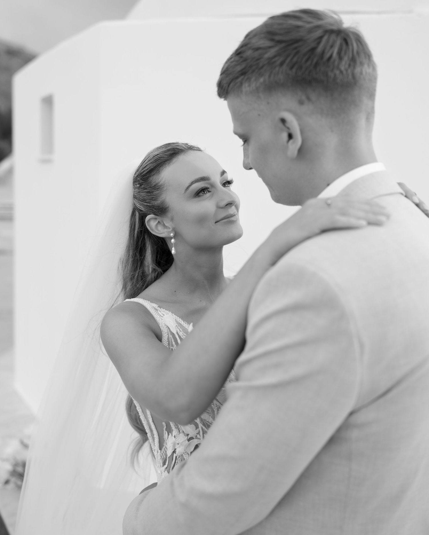 A romantic black and white portrait of a couple in wedding attire embracing intimately against a white wall.