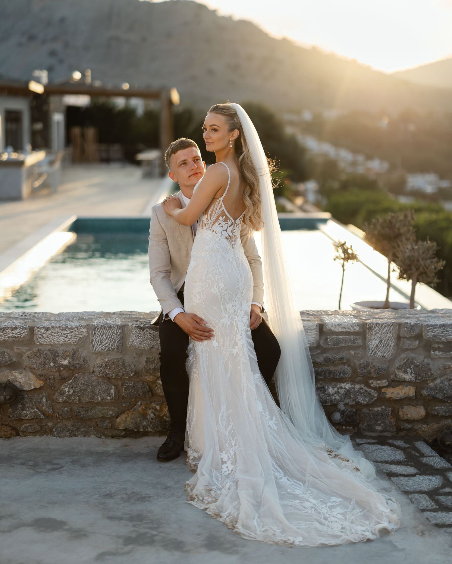 Romantic wedding couple embraces at sunset by luxury mountaintop pool with flowing lace dress and long veil.