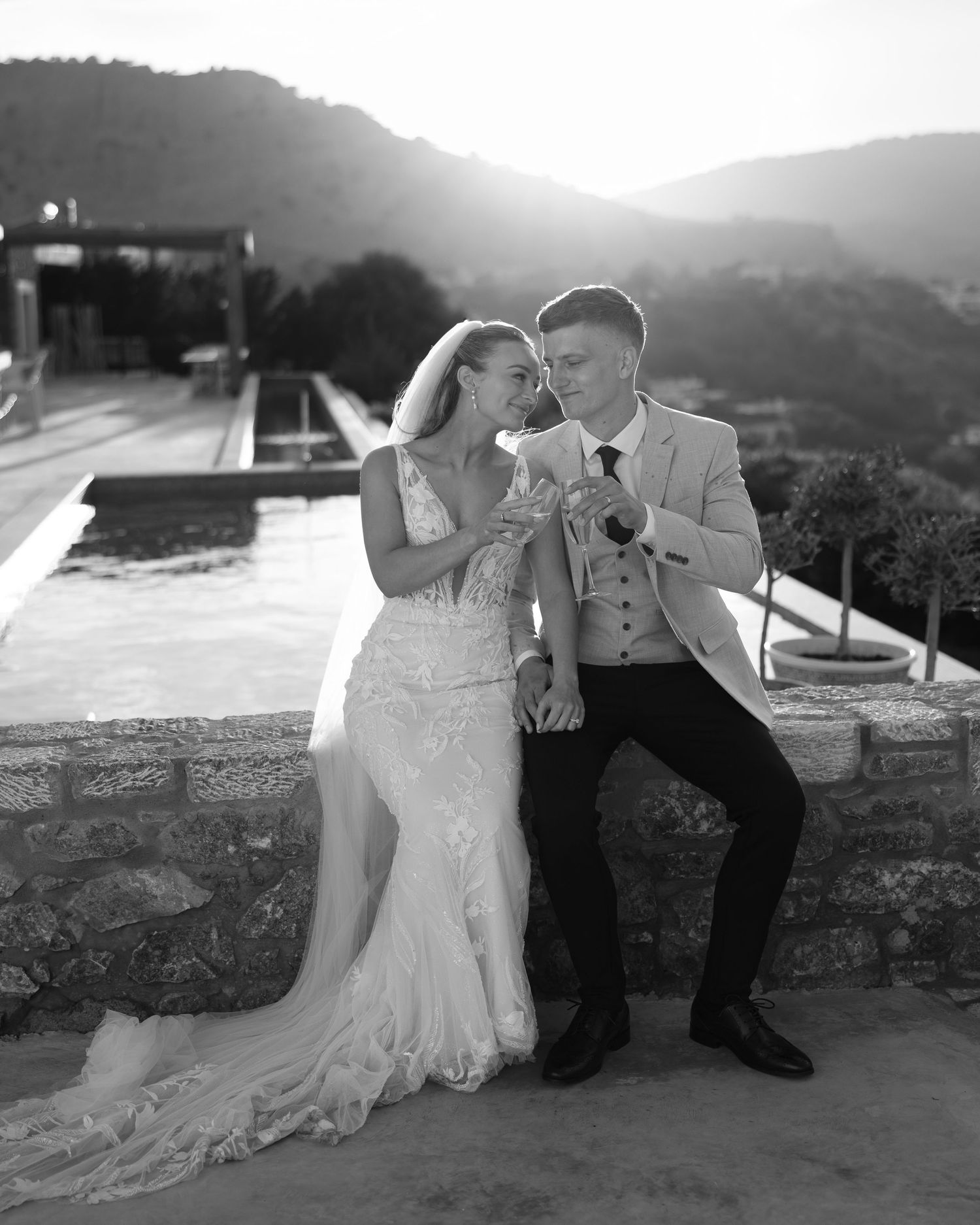 Newlywed couple shares romantic moment on stone terrace overlooking scenic mountain landscape at sunset.