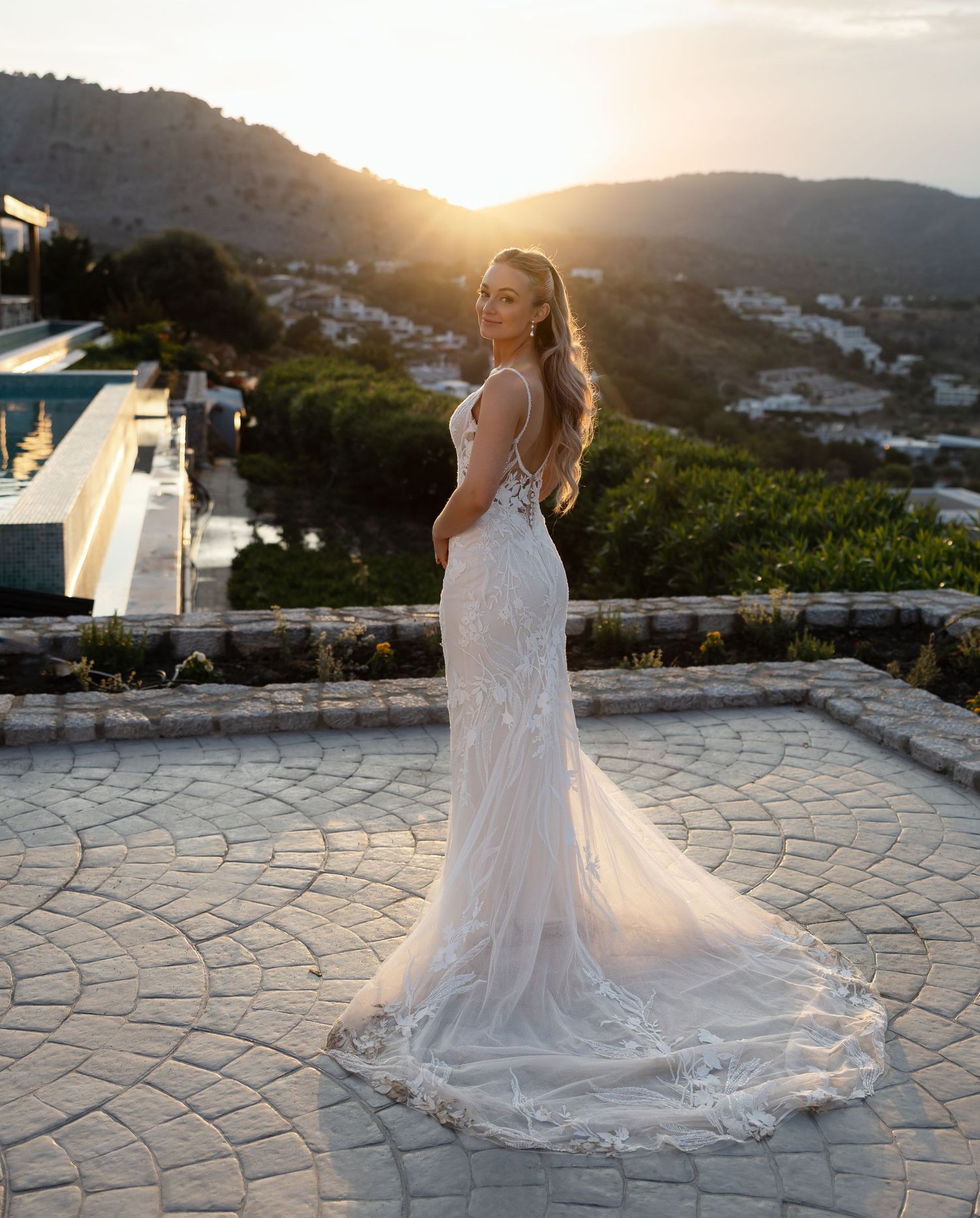 A bride in a flowing white wedding gown stands on a stone patio overlooking mountains at sunset in Greece.