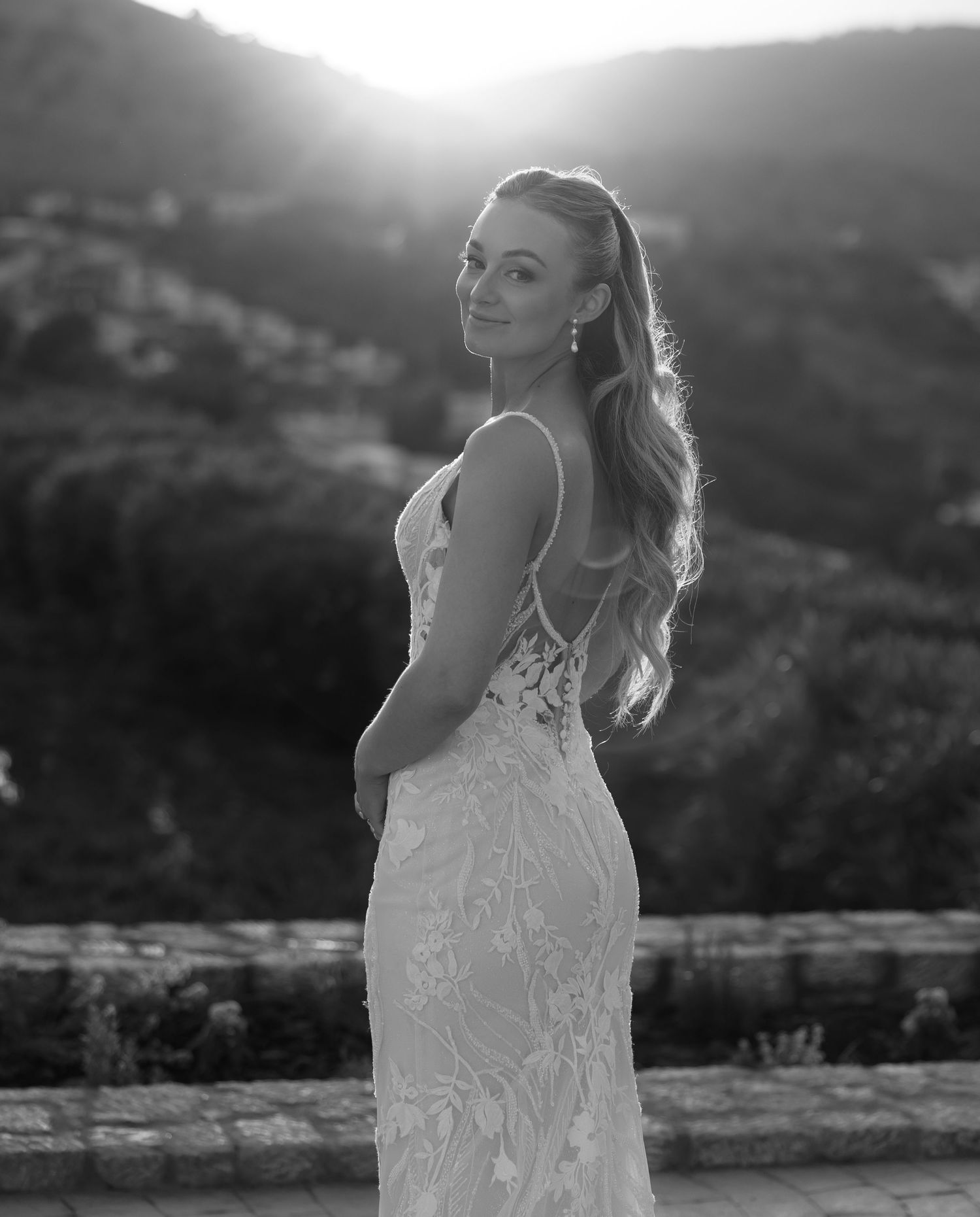 Black and white portrait of a bride in a lace wedding dress standing against a mountainous sunset backdrop.