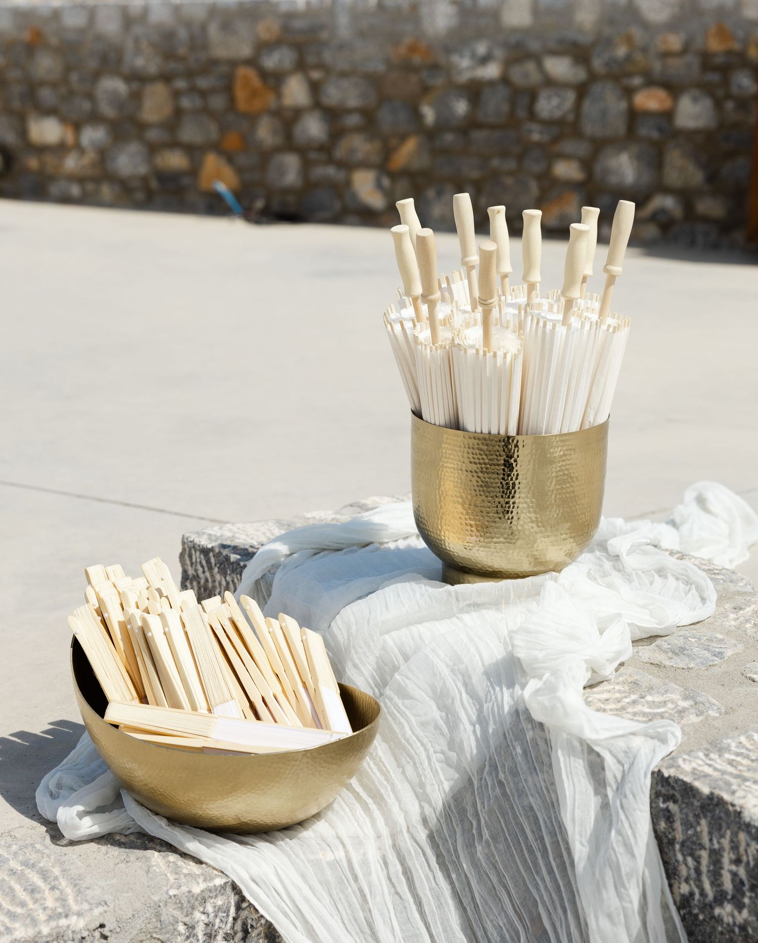 White straws and cocktail stirrers arranged in gold metallic containers on white tablecloth against stone wall background.