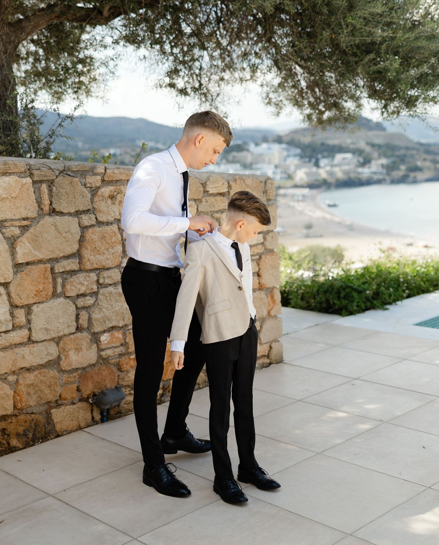 Two people in formal attire standing on a scenic coastal terrace with stone walls and mountain views in the background.