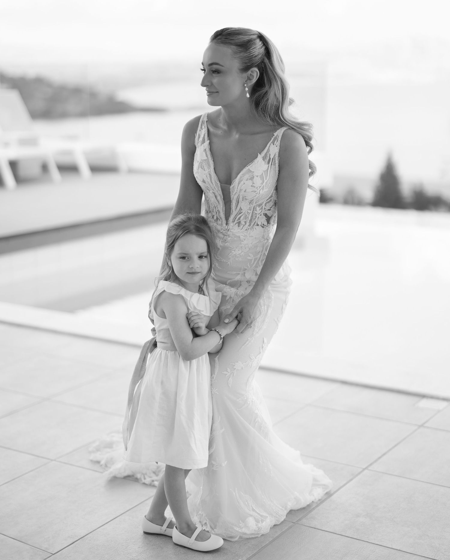 Black and white photograph of a bride in a lace wedding dress embracing a young flower girl in white on an outdoor terrace.