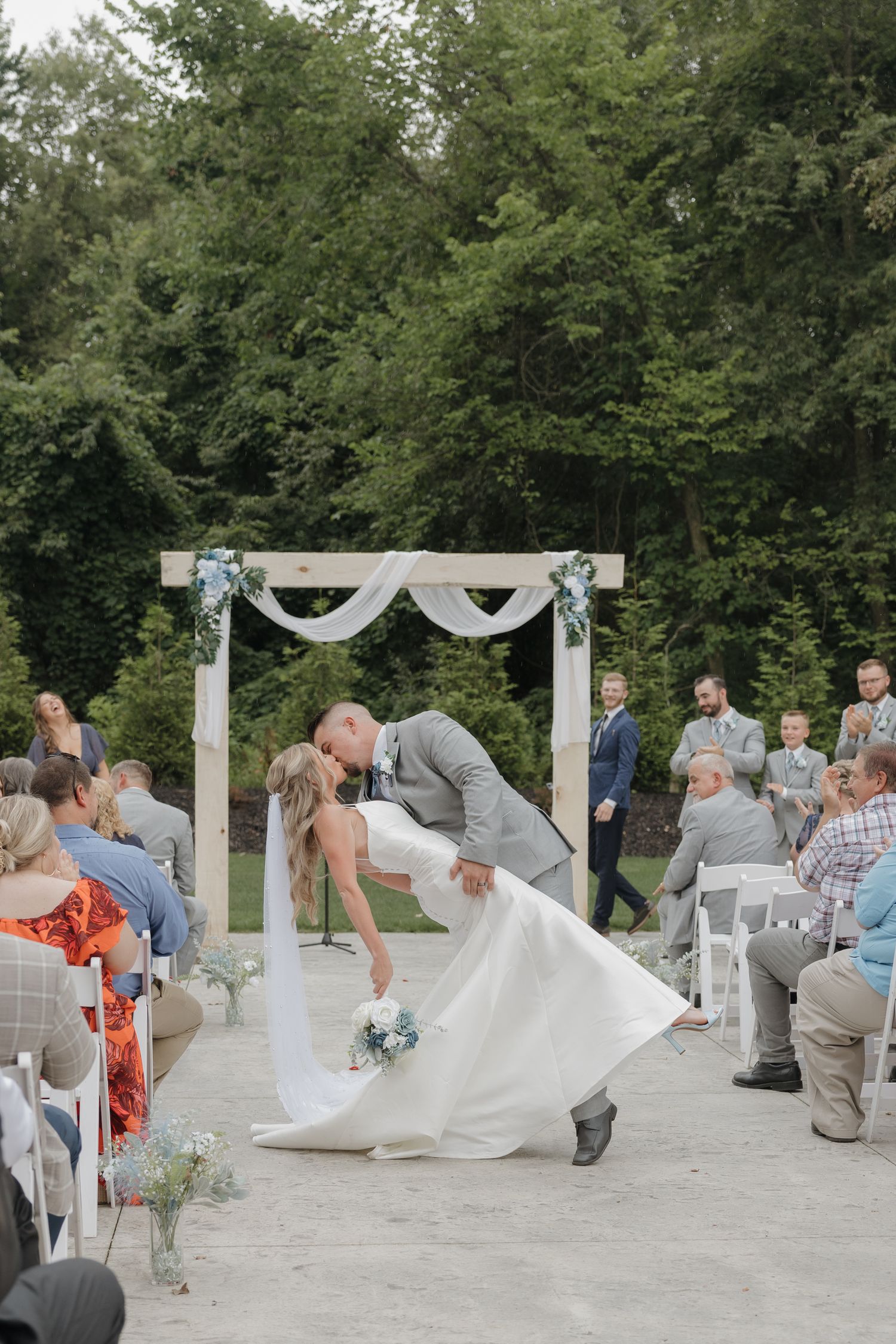 A romantic outdoor wedding ceremony shows a couple sharing their first kiss under a white draped arch surrounded by trees.