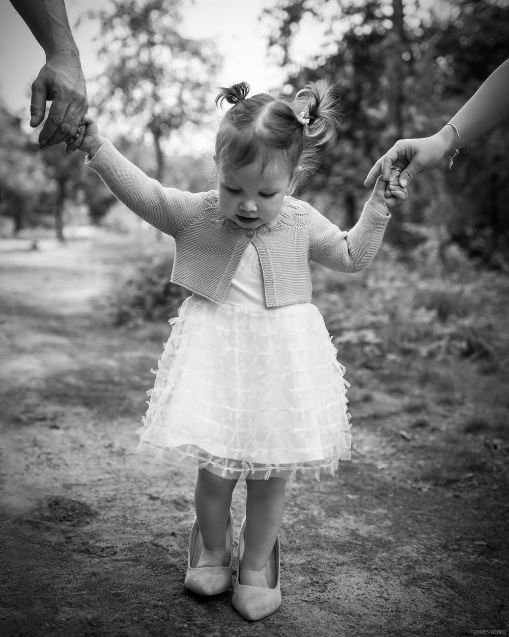 zwart wit foto van meisje die de te grote stiletto's van haar moeder draagt. ouders houder haar handjes vast. gefotografeerd voor verjaardag