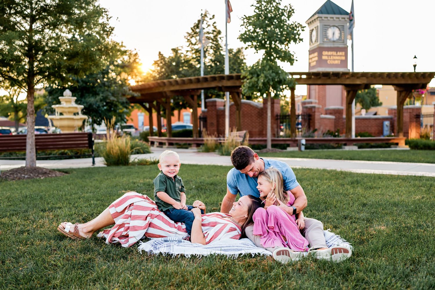 A family enjoys quality time together on a picnic blanket in a park with a clock tower in the background.